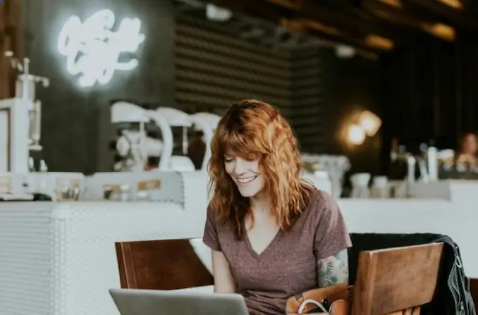 Young woman with  red hair wearing a purple sweater sitting at a wooden table in a dispensary, smiling while using a laptop computer in a bright, welcoming retail environment with white brick walls and warm lighting